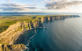 Aerial view of dramatic coastal cliffs rising above the Atlantic Ocean, with layered rock faces, green fields stretching across the cliff tops, and gentle waves below. Sunlight breaks through scattered clouds, illuminating the rugged coastline and patchwork farmland extending inland.