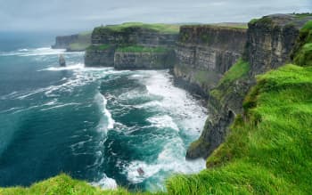 View of towering sea cliffs rising above the Atlantic Ocean, with steep rock faces topped by lush green grass. White waves crash against the shoreline below, and a solitary sea stack stands offshore beneath an overcast sky, creating a dramatic coastal landscape.