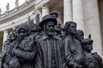 Bronze sculpture depicting a diverse group of people standing closely together, including figures wearing different styles of clothing and headwear. The monument is shown in the foreground with detailed facial expressions, while a large stone building with tall classical columns rises in the background.